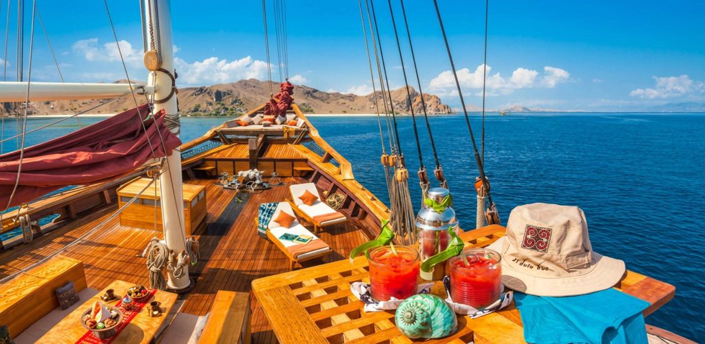 A luxury wooden phinisi yacht sailing through the crystal-clear blue waters of a tropical archipelago, featuring a sun-drenched deck set with tropical red cocktails, lounge chairs, and a panoramic view of remote, rolling desert islands.