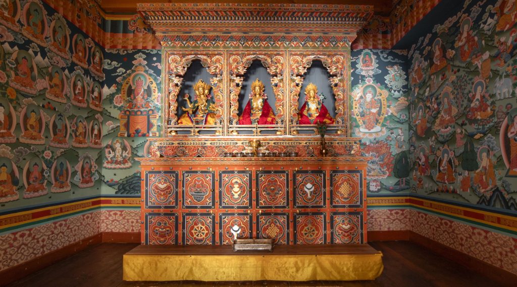 A Buddhist shrine inside a Bhutanese temple featuring three gilded Buddha statues on an intricately painted wooden altar surrounded by wall murals.
