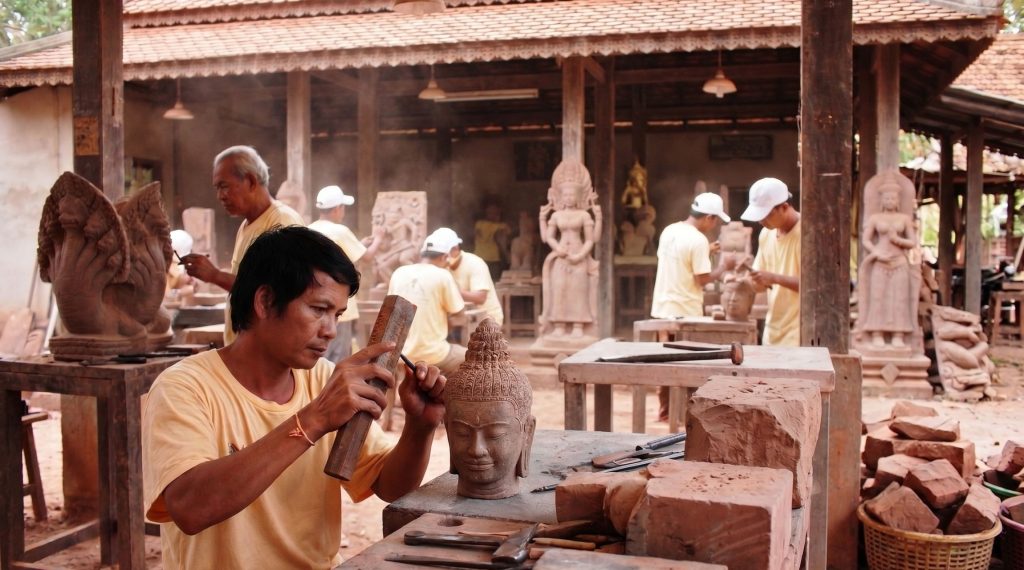 Traditional Cambodian stone carving workshop featuring a skilled artisan hand-carving a sandstone Buddha head with a mallet and chisel, surrounded by other sculptors.
