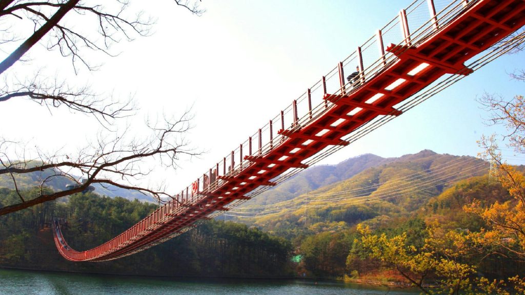 Low-angle view of a long, red pedestrian suspension bridge spanning across a calm lake, surrounded by lush green mountains and trees.