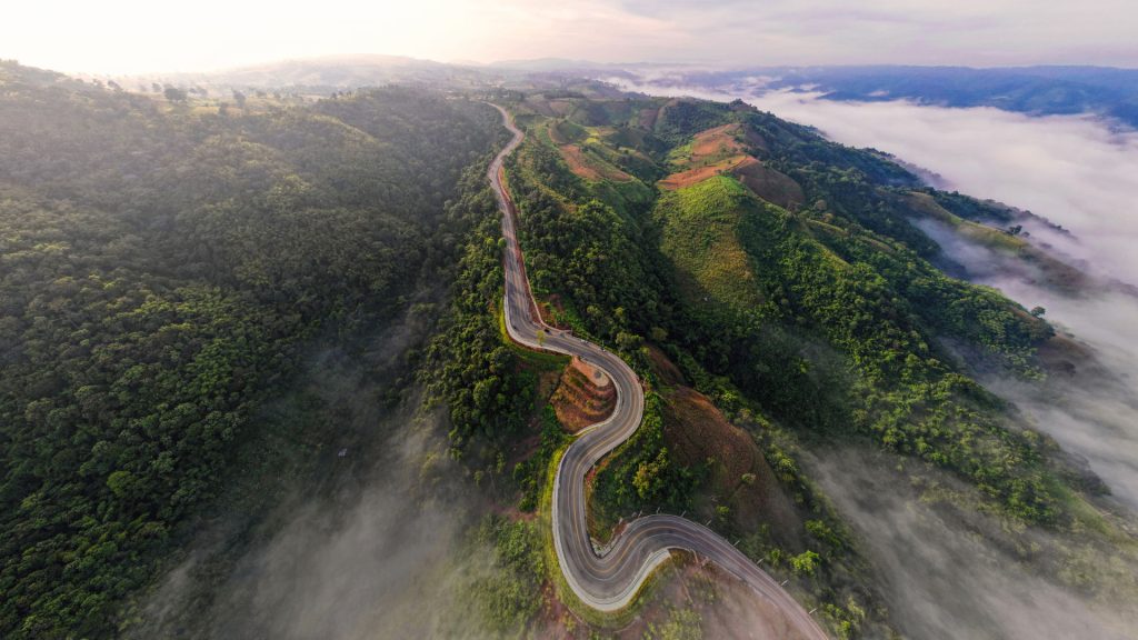 High-angle aerial view of a winding mountain road snaking through dense green hills shrouded in low-lying white clouds and morning mist.