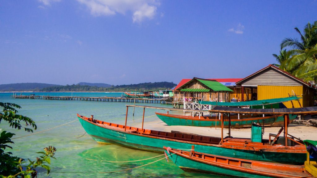 Colorful wooden fishing boats moored along a tropical sandy beach with shallow turquoise water and beachfront wooden huts.
