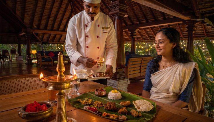 A photograph capturing a woman seated at a wooden dining table, with a waiter standing next to her, lighting a large brass traditional Indian oil lamp (diya). The waiter, dressed in a white uniform, holds a smaller flame to light one of the multiple wicks of the main kuthu vilakku stand. The woman, with a gentle smile and wearing a traditional Kerala kasavu saree and glasses, watches the lighting process. In the background, there is a spread of South Indian Sadhya food arranged on a banana leaf, a traditional uruli bowl with hibiscus flowers, and the wooden architecture and thatched roof of an open-air Kerala resort restaurant pavilion.