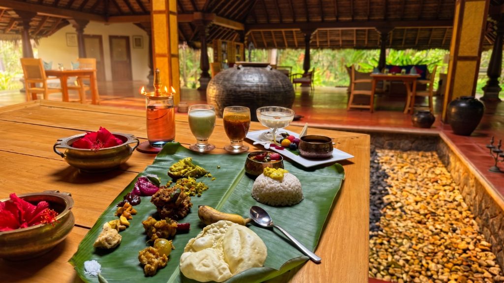 A close-up photograph capturing a traditional South Indian Sadhya meal, presented on a large, vibrant green banana leaf over a warm wooden table. The spread features a mound of white rice topped with yellow dal, and various colorful curries, side dishes, and condiments neatly arranged around it, including red pickles, mixed vegetable stir-fries, coconut chutneys, a small banana, and deep-fried papadums. In the background, there is a large, glowing brass oil lamp (kuthu vilakku) on a wooden stand, glasses containing different drinks, including a glass of water and a red drink, and a traditional uruli bowl filled with red hibiscus flowers, all within a traditional, open-air Kerala-style restaurant pavilion.
