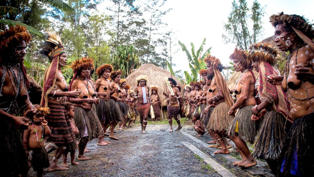 Indigenous villagers in traditional grass skirts, headdresses, and body paint gathering in an open dirt clearing in front of thatched huts.