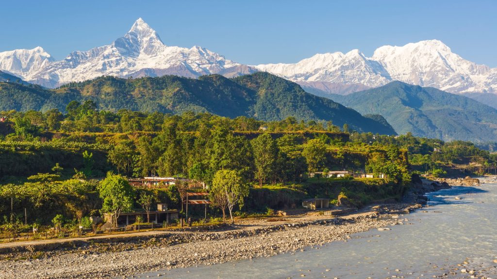 A wide, stony riverbed flowing past a green village valley with snow-capped Himalayan mountain peaks rising in the background under a clear sky.