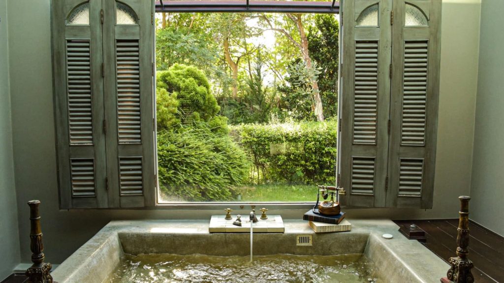 Rustic stone soaking bathtub overlooking a lush green garden through open louvered windows at a mountain retreat