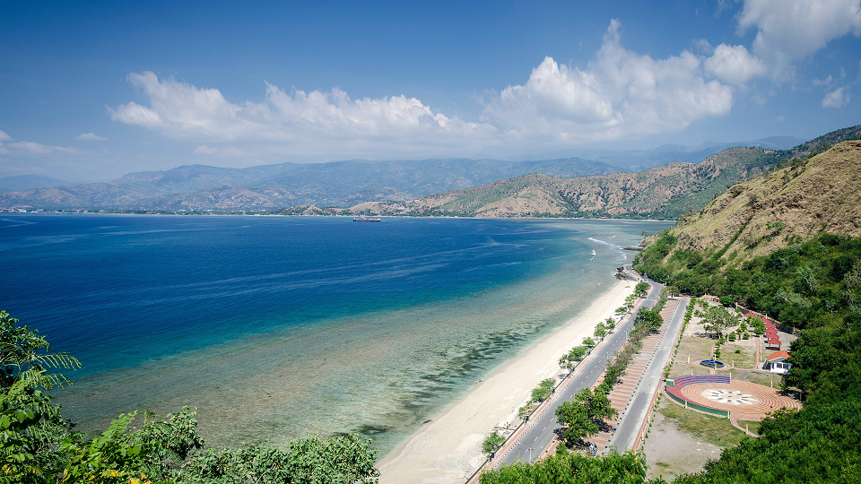 High-angle panoramic view of a crescent-shaped sandy coastline with turquoise water, lined by a paved beachfront road, a circular plaza, and dry, hilly terrain under a clear blue sky.