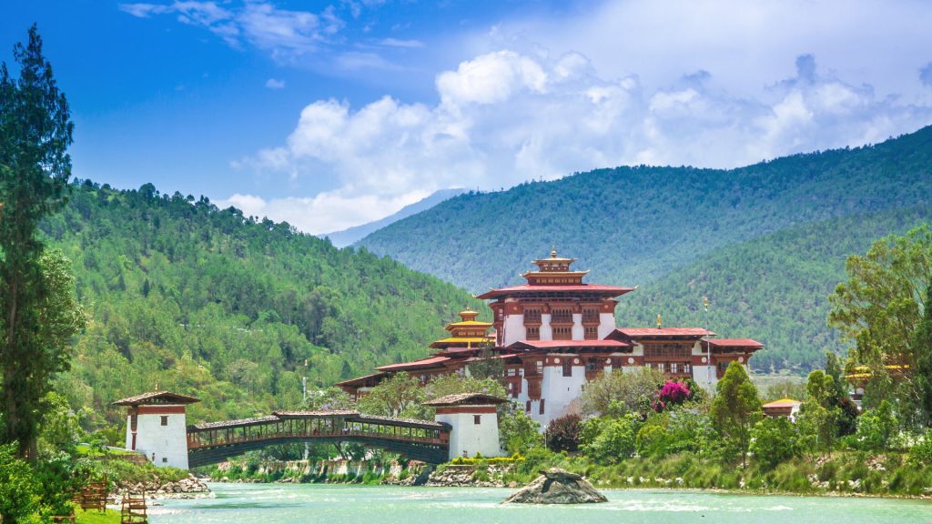 Traditional Bhutanese cantilever bridge leading to Punakha Dzong framed by forested Himalayan hills and a flowing river